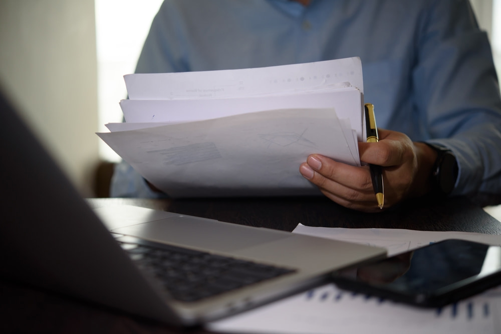 Person reviewing a stack of documents at a desk with a laptop, smartphone, and pen