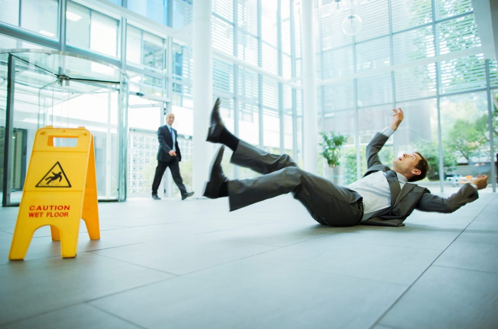 businessman falling backward on a wet floor beside a caution sign in an office lobby